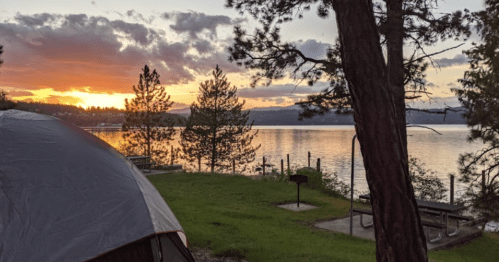 A serene lakeside view at sunset, with a tent in the foreground and trees framing the peaceful scene.
