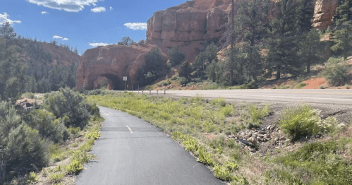 A scenic view of a paved path alongside a road, with red rock formations and greenery in the background.