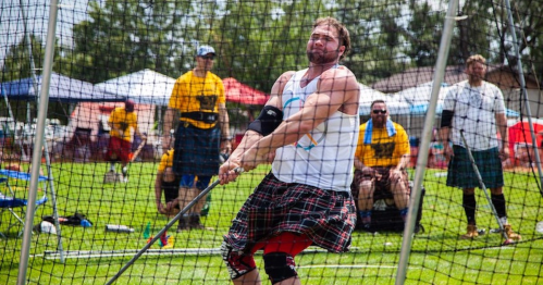 A man in a kilt swings a heavy weight in a sports competition, with spectators and tents in the background.