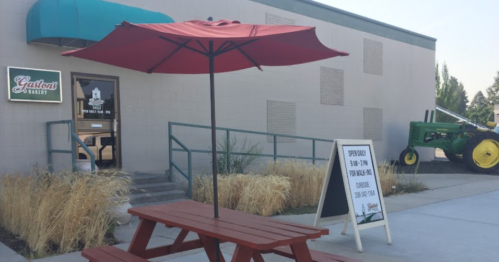 A red umbrella over a picnic table outside a bakery, with a green tractor and a sign nearby.
