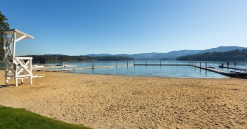A serene lakeside view with a sandy beach, docks, and mountains in the background under a clear blue sky.