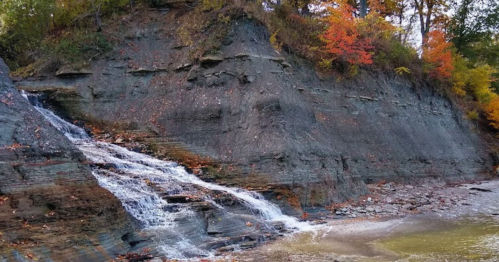 A small waterfall cascades down a rocky cliff, surrounded by autumn foliage in vibrant colors.