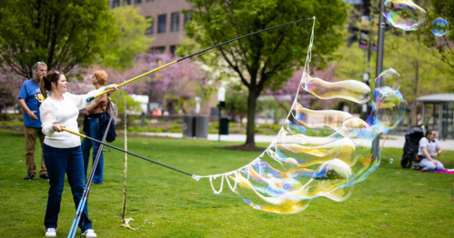 A woman creates large soap bubbles in a park, with people enjoying the scenery in the background.