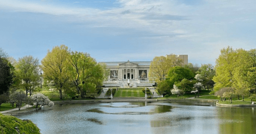 A serene view of a grand building surrounded by lush trees and a calm pond under a clear blue sky.