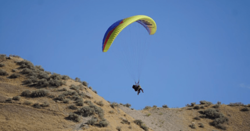 A person paragliding over a hilly landscape with sparse vegetation under a clear blue sky.