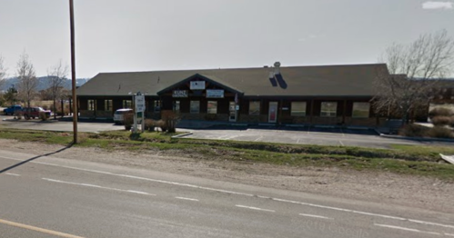A single-story building with a sloped roof, surrounded by grass, located along a road.