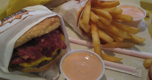 A close-up of a cheeseburger with corned beef, fries, and two cups of dipping sauce on a tray.