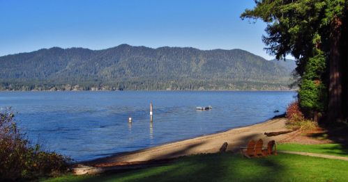 A serene lakeside view with mountains in the background, featuring a sandy shore and wooden chairs.