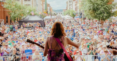 A musician performs on stage for a large crowd at an outdoor festival, with trees and buildings in the background.
