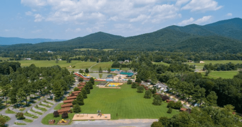 Aerial view of a park with playgrounds, pools, and green fields, surrounded by mountains and trees.