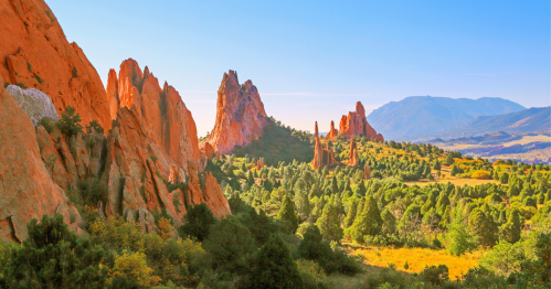 Vibrant red rock formations rise above a lush green landscape under a clear blue sky, with distant mountains in the background.