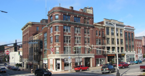 Historic brick buildings line a street, featuring shops and vehicles parked along the curb under a clear blue sky.