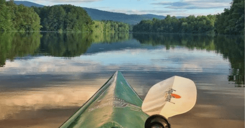 A kayak on calm water, reflecting trees and mountains under a clear sky. Paddle rests beside the kayak.