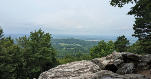A scenic view from a rocky outcrop, overlooking lush green hills and a cloudy sky in the distance.