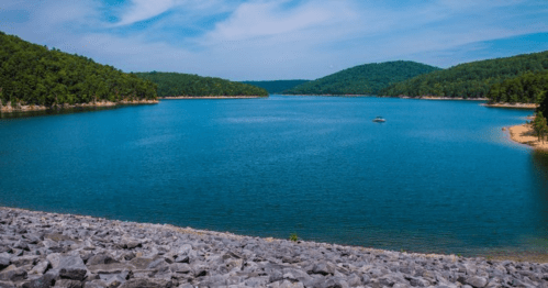 A serene lake surrounded by green hills under a clear blue sky, with a rocky shoreline in the foreground.