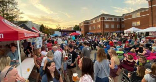 A lively outdoor event with a large crowd enjoying music, food, and drinks under a sunset sky.