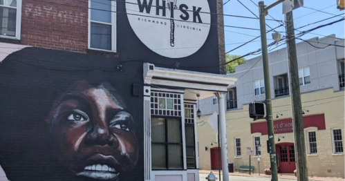 A mural of a woman's face on a brick wall beside a sign for Whisk in Richmond, Virginia. Power lines overhead.