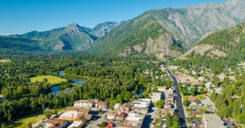 Aerial view of a small town surrounded by mountains and greenery, with a river flowing nearby. Clear blue sky above.