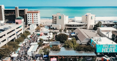 Aerial view of a bustling beachside area with shops, tents, and a clear blue ocean in the background.