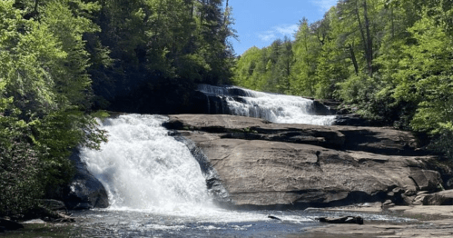A serene waterfall cascades over rocky ledges, surrounded by lush green trees under a clear blue sky.