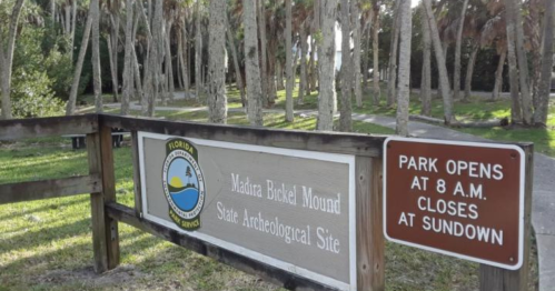 Sign for Madira Bickel Mound State Archeological Site, with park hours displayed, surrounded by palm trees.