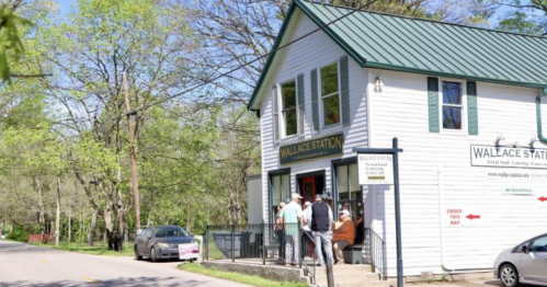 A quaint white building with a green roof, labeled "Wallace Station," surrounded by trees and a road. People are gathered outside.