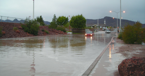 A flooded road with a car navigating through standing water, surrounded by trees and mountains in the background.