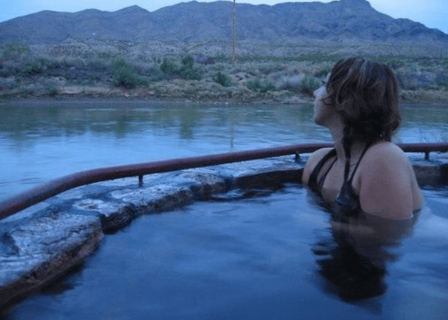 A person relaxes in a hot spring, gazing at mountains under a twilight sky.