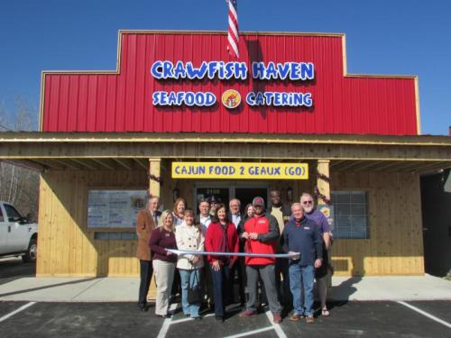 A group of people stands outside Crawfish Haven, a seafood catering business, holding a ribbon for a grand opening.