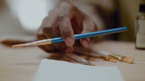 A close-up of a hand holding a blue paintbrush, surrounded by various art supplies on a wooden surface.