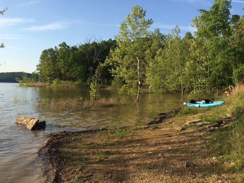 A serene lakeside scene with a kayak on the shore, surrounded by lush green trees and calm water under a clear sky.