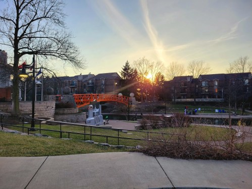 Sunset over a park with an orange bridge, trees, and apartment buildings in the background.