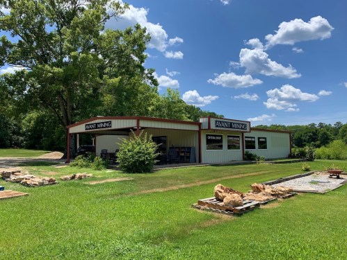 A mining shop with a red and white exterior, surrounded by green grass and trees, under a blue sky with fluffy clouds.