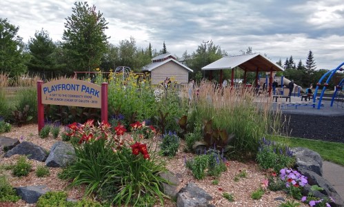 A park sign reads "Playfront Park," surrounded by colorful flowers, with playground equipment and a shelter in the background.