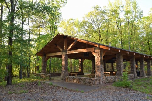 A rustic pavilion with stone pillars surrounded by lush green trees in a serene outdoor setting.