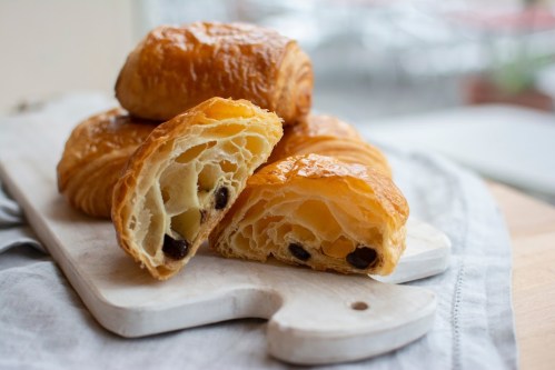 A stack of flaky croissants on a wooden board, with one cut open to reveal chocolate filling inside.