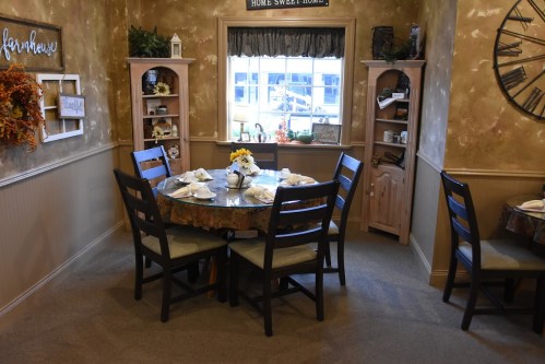 Cozy dining area with a round table set for four, surrounded by warm decor and shelves with decorative items.