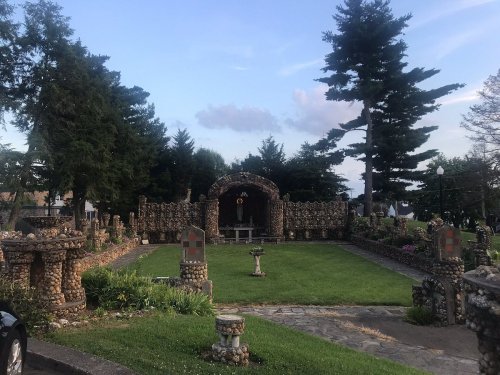 A stone garden with a central archway, surrounded by trees and decorative stone structures, under a blue sky.