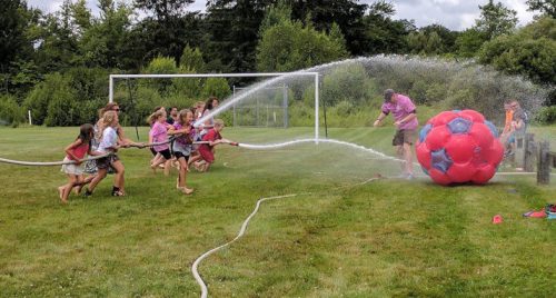 Children play with a large inflatable ball while spraying water from hoses on a grassy field.