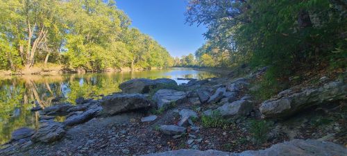 A serene river scene with rocky banks, surrounded by lush green trees and clear blue skies reflecting on the water.