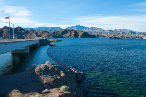 A bridge spans a blue lake surrounded by mountains under a clear sky, with a flag waving in the foreground.