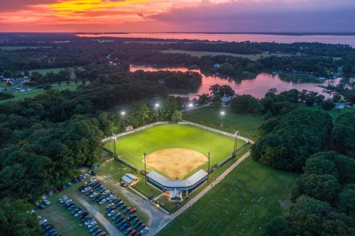 Aerial view of a baseball field at sunset, surrounded by trees and a lake, with cars parked nearby.