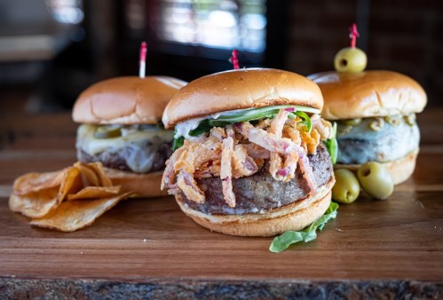 Three gourmet burgers on a wooden board, topped with greens, crispy onions, and served with potato chips and olives.