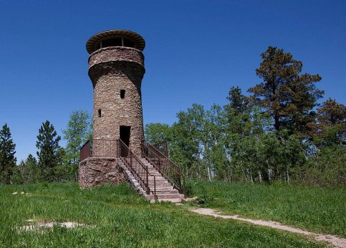 A stone observation tower with a staircase, surrounded by green grass and trees under a clear blue sky.