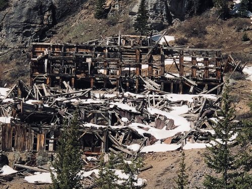 Abandoned wooden structure in a mountainous area, partially covered in snow and surrounded by trees.