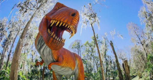 A large, colorful dinosaur statue stands in a forested area with trees and Spanish moss in the background.