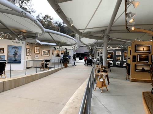 A gallery walkway with art displays, featuring framed photos and paintings under a modern canopy. Visitors are exploring.
