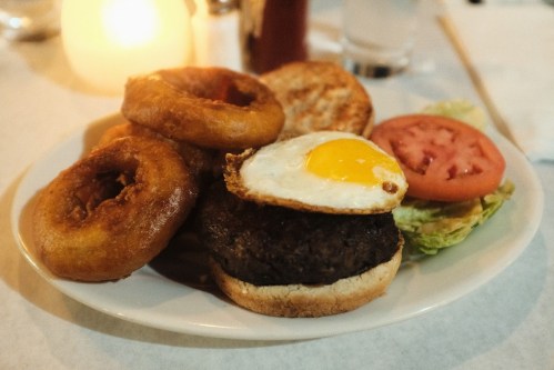 A plate with a burger topped with a fried egg, onion rings, a slice of tomato, and lettuce on a table.