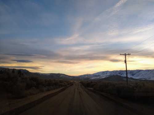 A dirt road stretches into the distance, flanked by mountains and a colorful sunset sky with wispy clouds.