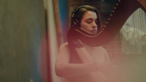 A musician plays a harp, wearing headphones, with a soft focus background.